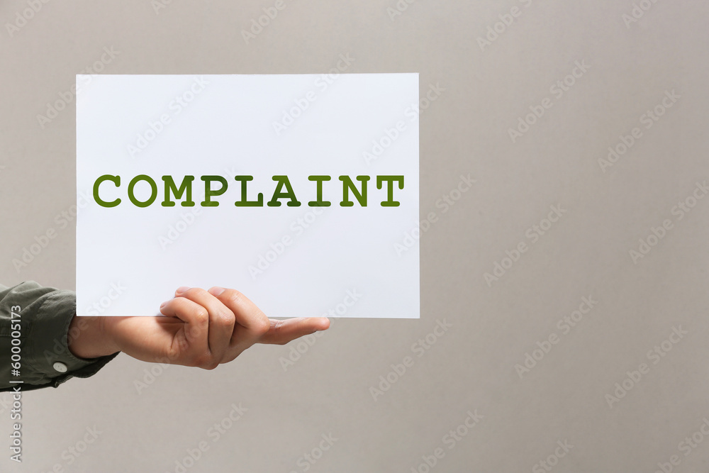 Man holding sheet of paper with word Complaint on grey background, closeup. Space for text Man holding sheet of paper with word Complaint on grey background, closeup. Space for text