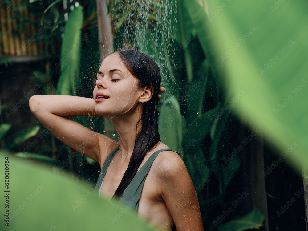 A woman in a swimsuit washes her head in a tropical shower outdoors against the backdrop of green tropical leaves, flowers and palm trees. Body and hair care, tanned skin, sexy, open mouth - obrazy, fototapety, plakaty 