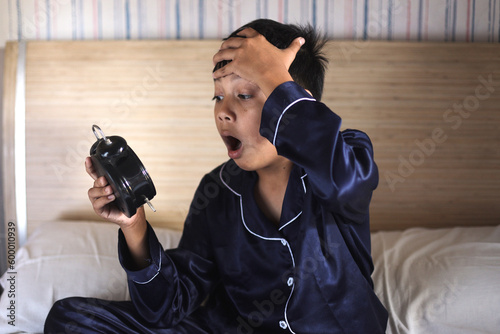 Worried awake teen boy with alarm clock is shocked because it is too late
