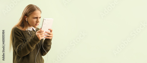 Canvas Print Frowning little girl in earphones using tablet computer on light green backgroun
