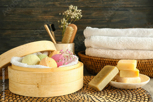 Box with bath bombs, flowers, soap bars and towels on mat near dark wooden wall, closeup