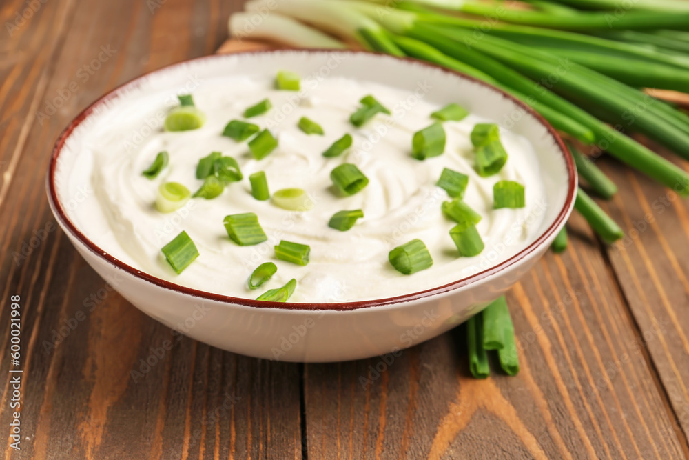 Bowl of tasty sour cream with green onion on wooden background, closeup