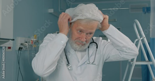 old experienced senior doctor putting on cap, hat in the hospital, preparaing for surgery, operation Portrait of surgeon in uniform and safety mask putting on hat going to work with patients photo