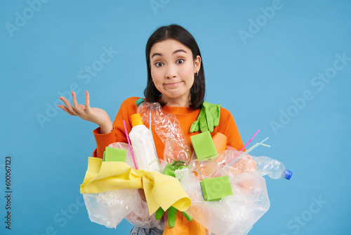 Korean girl with plastic garbage, shrugs shoulders, doesnt know how to recycle, sorting her waste for recycling station, blue background photo