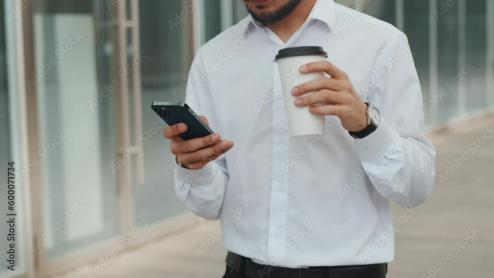 Young businessman in a white shirt walks near the business center with a smartphone and drinking coffee from a paper cup outdoors, lunch break Young businessman in a white shirt walks near the business center with a smartphone and drinking coffee from a paper cup outdoors, lunch break
