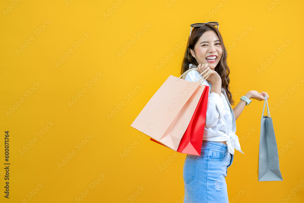 Fototapeta premium cheerful shopper smiling and holding shopping bags isolated on yellow studio background, happy young Asian pretty woman enjoy purchasing gifts, for advertising and sale promotion shopaholic concept