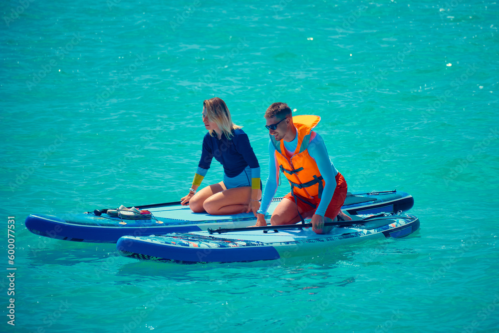 Fototapeta premium Summer outdoor seaside activities. Man and woman swim on supboards. Young man dressed in swimsuit and lifevest and young woman swimming on stand up paddle board.