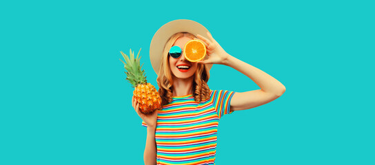 Summer portrait of happy smiling young woman with pineapple and slice of orange, fresh tropical juicy fruits, wearing straw hat, sunglasses on blue background
