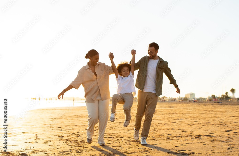 Seaside serenity concept. Happy multiracial family walking along coastline, parents lifting son up, having fun outdoors