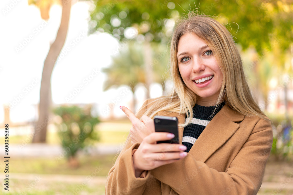 Young pretty blonde woman at outdoors using mobile phone and pointing back Young pretty blonde woman at outdoors using mobile phone and pointing back