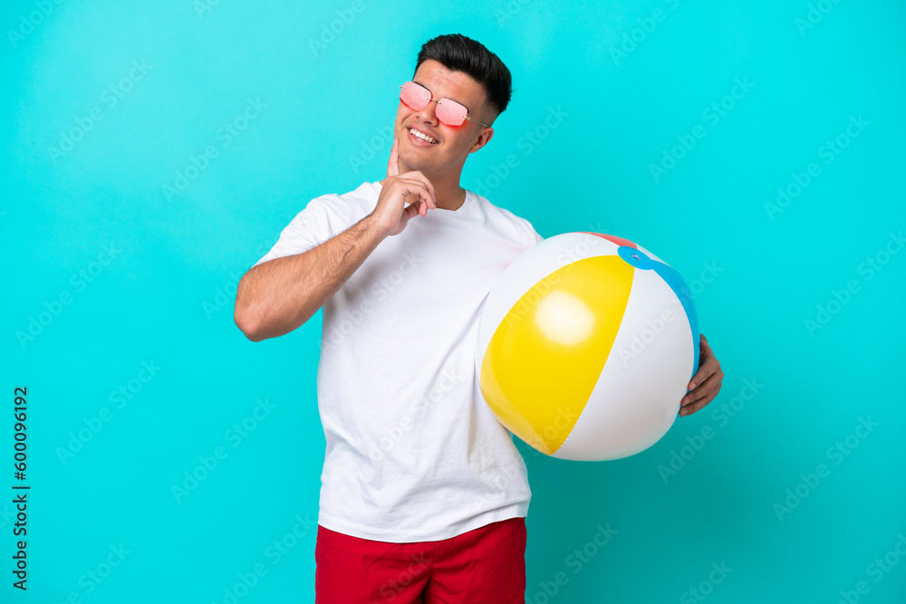 Young caucasian man holding a beach ball isolated on blue background thinking an idea while looking up