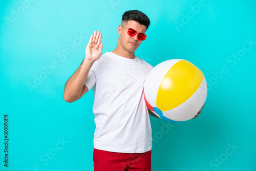 Young caucasian man holding a beach ball isolated on blue background making stop gesture and disappointed