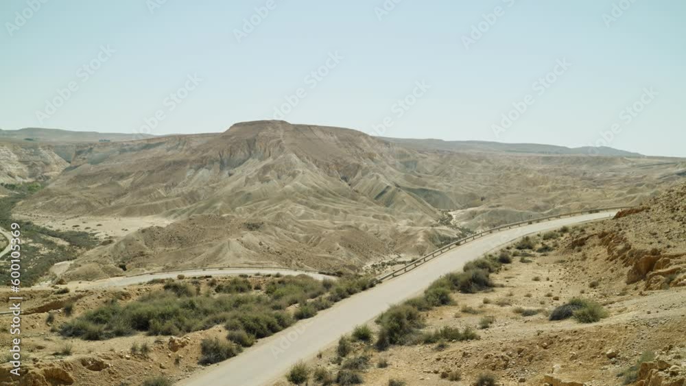 Empty desert winding road on hot sunny day during summer with mountain and dunes. Green vegetation. Small back country road or off-road going down to bare and desolate valley between sandy hills