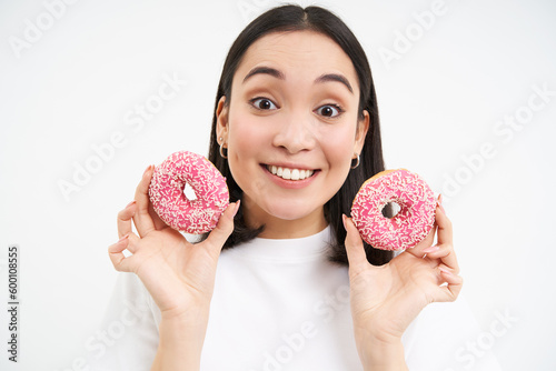 Smiling korean woman eats pink glazed doughnuts, enjoys delicious donnut, junk food, not care about diet, white background photo