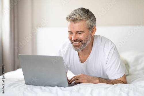 Relaxed mature man lying on bed, using laptop, copy space