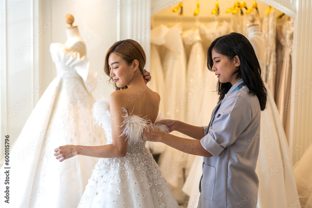 Portrait of smiling happy love asian fashion bride woman talk and try on wedding dress with woman shop owner for pre wedding at wedding interior fashion studio.