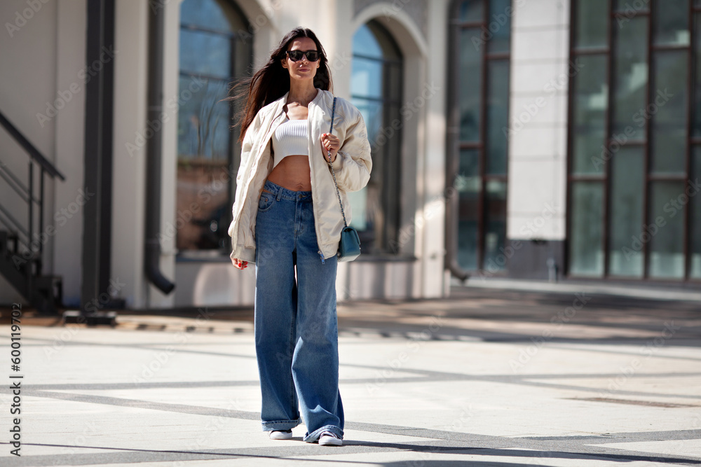 Fashionable woman with long flowing hair wearing trendy white bomber jacket, short Tshirt, wide jeans, posing on street of city. Trends clothing of spring and summer