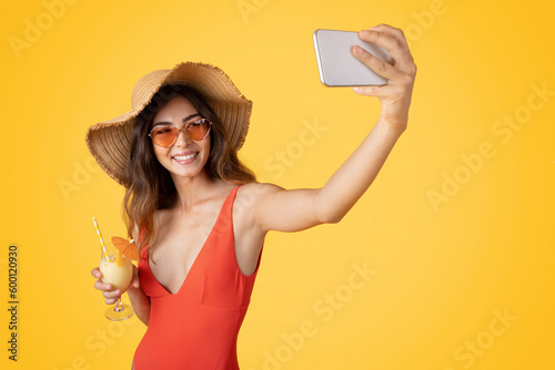 Smiling millennial asian woman in swimsuit, sunglasses and hat with tropical cocktail, enjoy free time #600120930