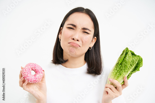 Healthy clean detox eating concept. Vegetarian, vegan, raw concept. Sad asian girl on diet holds doughnut and vegetable, wants to taste donnut, white background photo