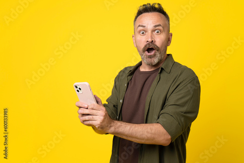 attractive amazed bristle man using digital smartphone in hands demonstrating shock from news or sms, reaction influencer post review isolated over yellow background