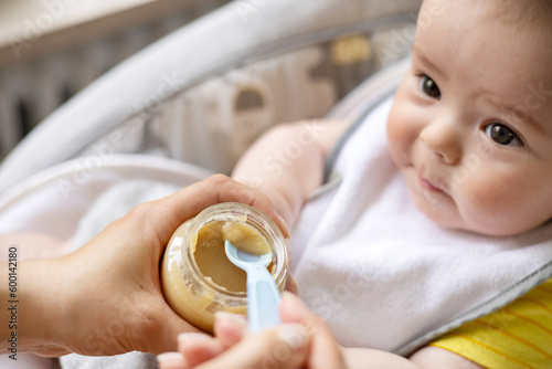 adorable cute baby boy sitting in swing cradle eating mashed food with spoon.mother hand feeding infant toddler.child with bib is smiling,no teeth on gum.vegetables or fruits puree in jar.hungry