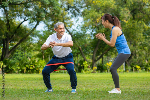 Senior asian man is using rubber band to build up his leg muscle strength while his daughter is cheering up in the public park for elder longevity exercise and outdoor workout usage