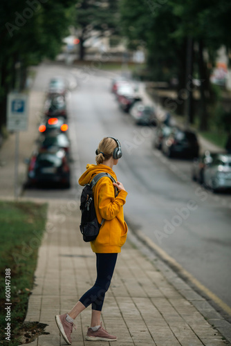 A woman with a city backpack and wearing headphones stands by the road. Back view.