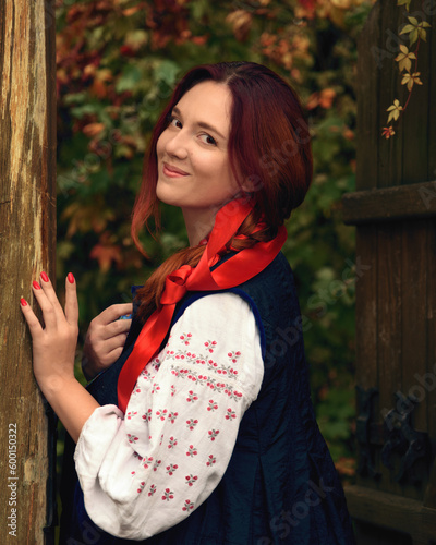 Smiling cheerful young redhead woman dressed traditional ukrainian embroidered clothes and with a red ribbon in her hair looking at the camera. Vintage outfit. Half-length portrait.