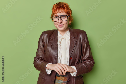 Portrait of goof looking ginger young womann with short hair dressed in leather jacket and whiye blouse looks sefl confident at camera isolated over green background ready for corporate meeting