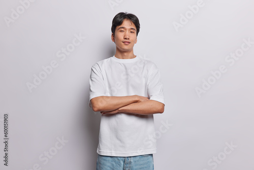 Horizontal shot of serious Asian man keeps arms folded stands in self assured pose looks confidently at camera dressed in casual t shirt and jeans isolated over white background. People and ethnicity