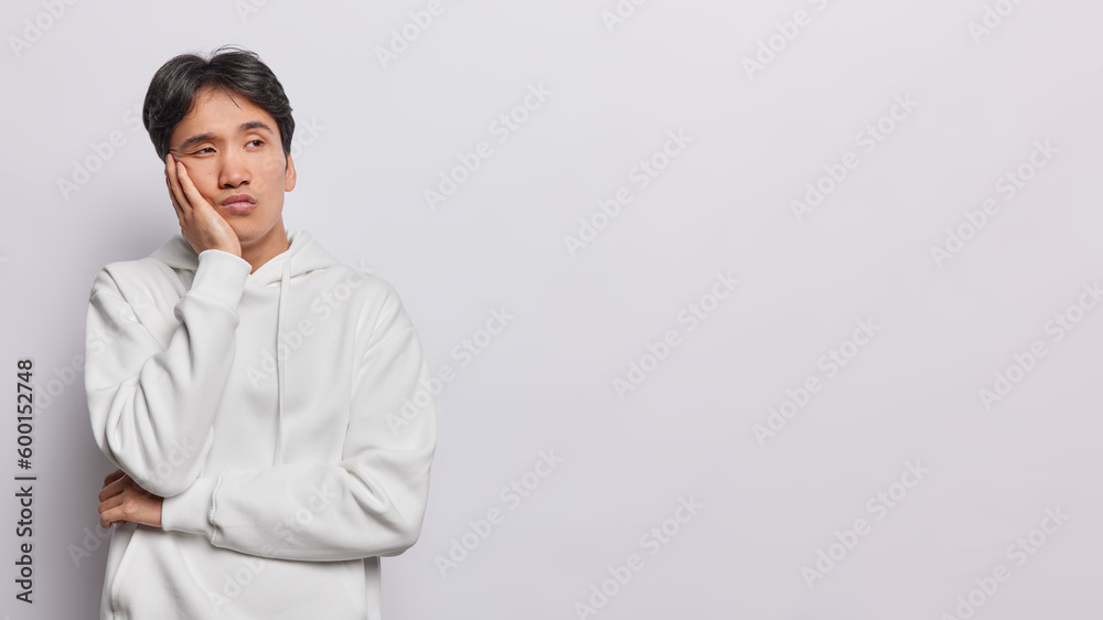 Studio shot of thoughtful puzzled Asian man keeps hand on cheek feels bored and unaware concentrated aside dressed in casual hoodie isolated over white background with copy space for your promotion Studio shot of thoughtful puzzled Asian man keeps hand on cheek feels bored and unaware concentrated aside dressed in casual hoodie isolated over white background with copy space for your promotion