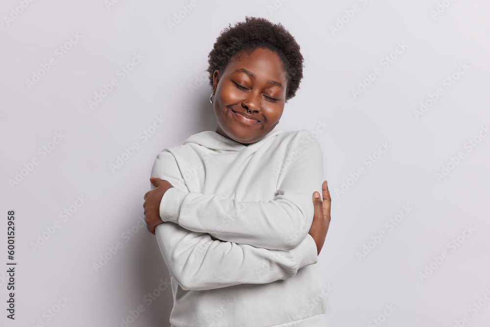 Body positive young woman embraces herself has good self esteem wears casual hoodie has eyes closed dreams about something isolated over white background. Body acceptance and self love concept - obrazy, fototapety, plakaty 