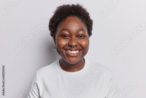 Portrait of chubby dark skinned woman smiles toothily being in good mood poses in studio for making photo wears earrings and piercing in nose isolated over white background. Happy emotions concept © Wayhome Studio