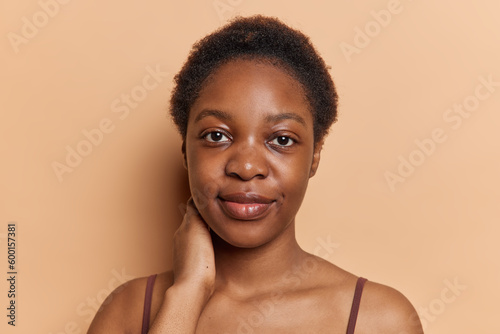 Portrait of teenage girl with dark skin short curly hair keeps hand on neck concentrated at camera stands with bare shoulders against brown background. Afro American woman looks seriously at camera