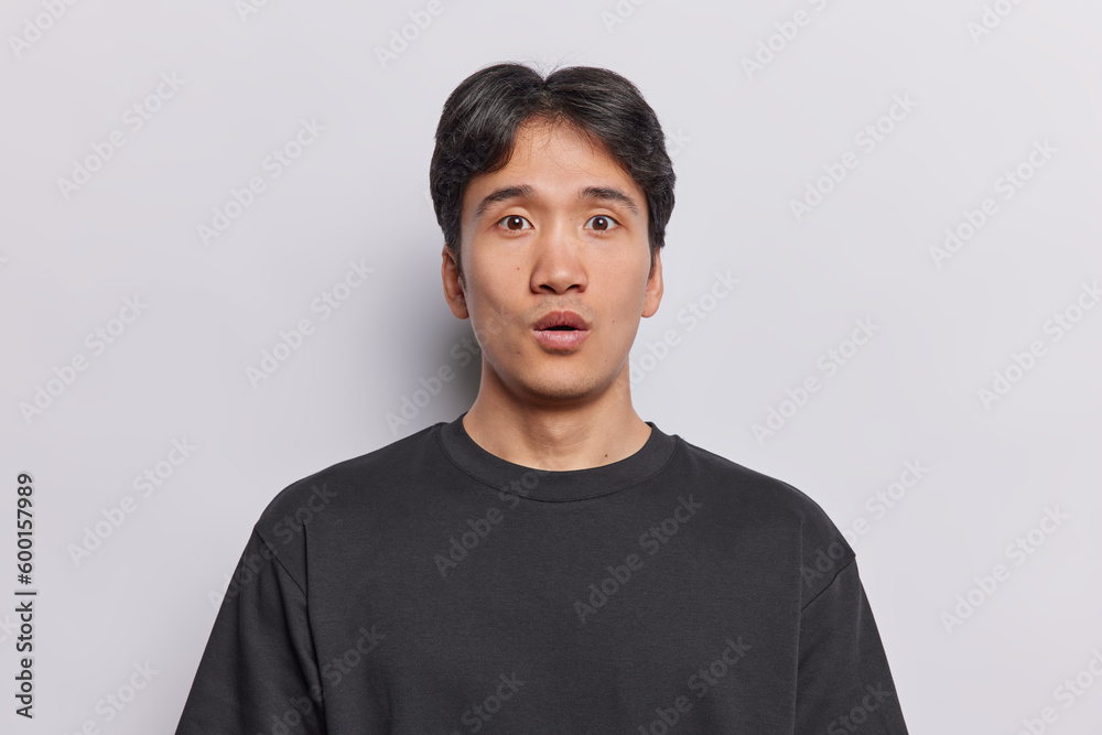 Surprised young Asian man with dark short hair feels amazed stands speechless reacts to shocking news dressed in casual black t shirt poses against white background. Human reactions concept.