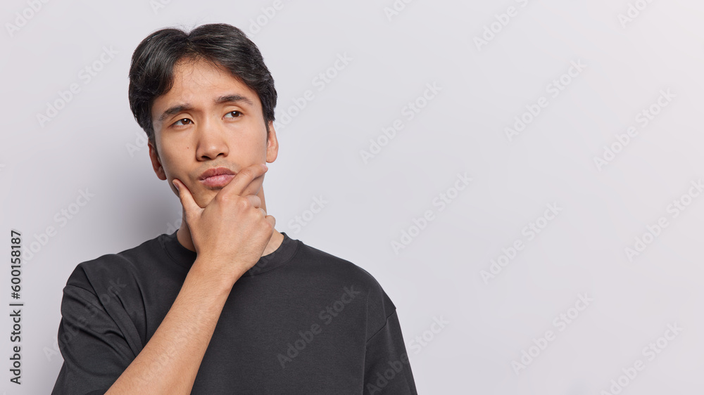 Horizontal shot of dark haired man keeps hand on chin looks thoughtfully aside ponders on something dressed in black t shirt isolated over white background copy space for your advertising content Horizontal shot of dark haired man keeps hand on chin looks thoughtfully aside ponders on something dressed in black t shirt isolated over white background copy space for your advertising content