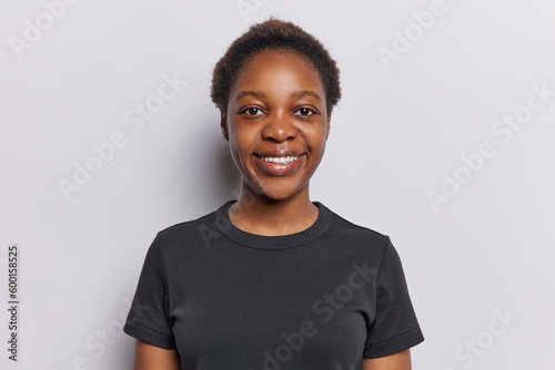 Canvastavla Portrait of charming dark skinned teenage girl looks at camera with smile expresses positiveness dressed in casual black t shirt isolated over white background
