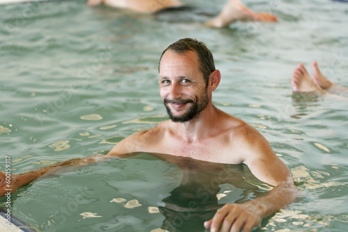 Happy mid adult, middle aged caucasian guy smiling in the indoor swimming pool with dense salt water, Dead Sea. Bearded man relaxing, enjoying in the spa pool. Positive delighted man sitting in water