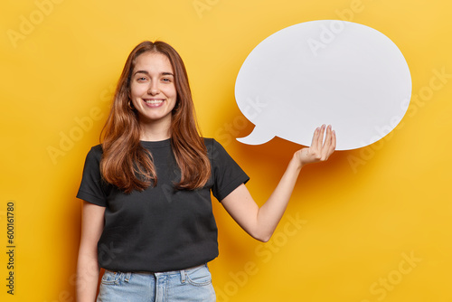 Waist up shot of satisfied millennial girl holds blank speech bubble smiles broadly suggests to place your advertising content here dressed in black t shirt and jeans isolated over yellow background