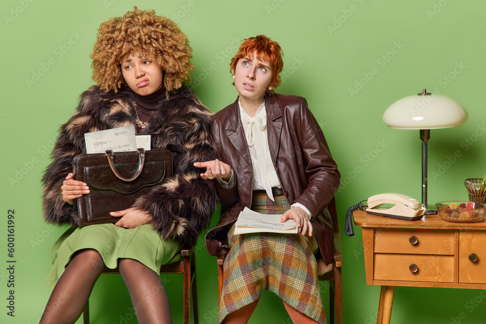 Two displeased young women dressed in retro clothes have clueless expressions sit on chairs hold papers being tired of waiting going to apply for work isolated over green background. Nostalgia concept