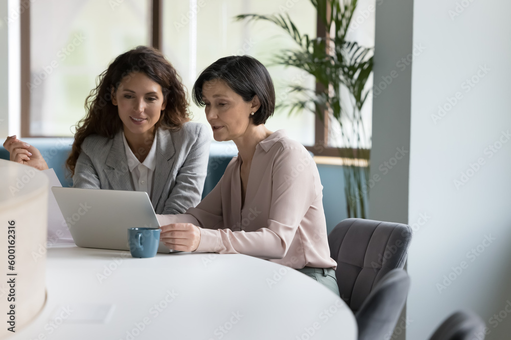 35s and 55s women colleagues working together use computer, meet in office staring at laptop screen, learn new software, younger Hispanic affiliate helps to older with program or app. Tech, business 35s and 55s women colleagues working together use computer, meet in office staring at laptop screen, learn new software, younger Hispanic affiliate helps to older with program or app. Tech, business
