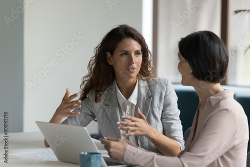 Foto Two business women sit at desk discuss project details, diverse female colleague