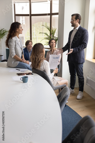 Confident businessman, male boss in suit explains new project details, share business strategy, makes speech stand in front of company staff members during corporate group briefing in conference room