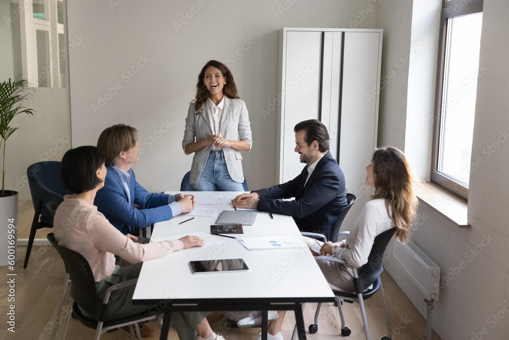 Group of cheerful businesspeople gather in conference room during morning briefing, take part in formal meeting, joking having friendly workflow in modern office, training led by Hispanic female coach Group of cheerful businesspeople gather in conference room during morning briefing, take part in formal meeting, joking having friendly workflow in modern office, training led by Hispanic female coach