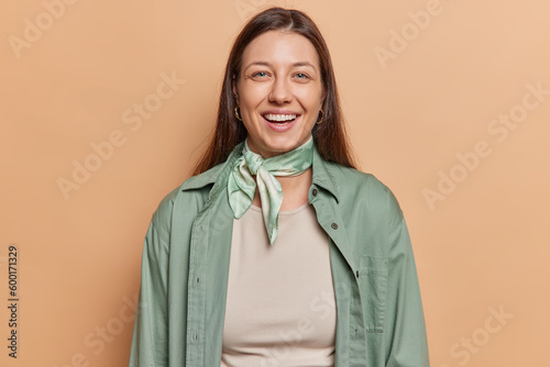 Portrait of attractive dark haired woman smiles happily wears green shirt and kerchief around neck being in good mood poses against brown background feels very positive. People and emotions concept
