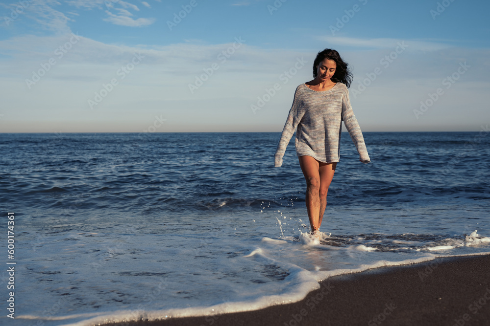 young beautiful brunette girl walks on the sand on the seashore in a swimsuit and a sweater young beautiful brunette girl walks on the sand on the seashore in a swimsuit and a sweater
