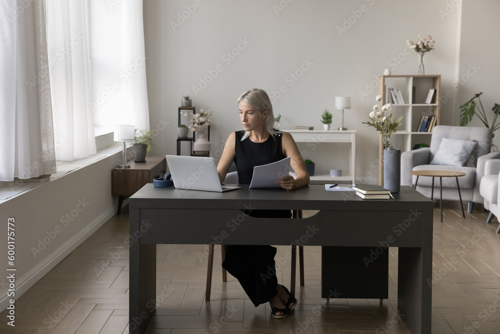 Confident mature business lady working from home, sitting at workplace table in living room, checking paper document, using online app, software for job communication. Full length shot
