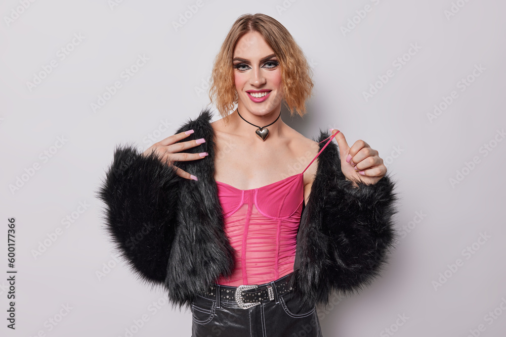 Horizontal shot of cheerful transgender man with short hair wears bright vivid makeup smiles toothily wears pink t shirt trousers and fur coat isolated over white background belongs to lgbt community Horizontal shot of cheerful transgender man with short hair wears bright vivid makeup smiles toothily wears pink t shirt trousers and fur coat isolated over white background belongs to lgbt community