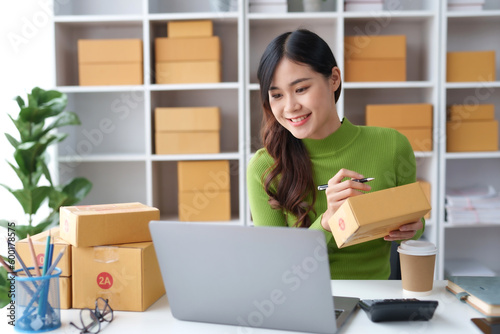 Smiling young Asian business owner woman prepare parcel box and check online orders of product for deliver to customer on laptop computer at home. Shopping Online concept. Startup small business SME. © amnaj