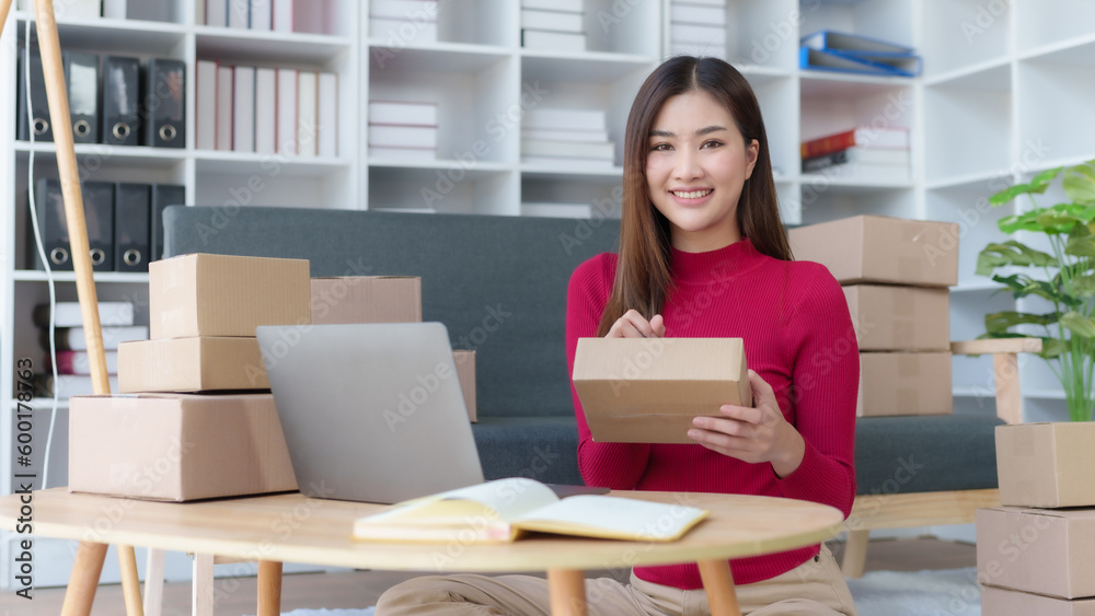 Smiling young Asian business owner woman prepare parcel box and check online orders of product for deliver to customer on laptop computer at home. Shopping Online concept. Startup small business SME. Smiling young Asian business owner woman prepare parcel box and check online orders of product for deliver to customer on laptop computer at home. Shopping Online concept. Startup small business SME.
